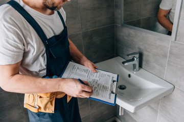 partial view of male plumber with toolbelt writing in clipboard near broken sink in bathroom