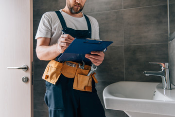 partial view of male plumber with toolbelt writing in clipboard near broken sink in bathroom