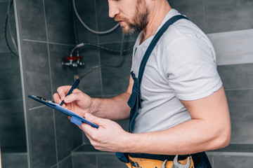 partial view of plumber writing in clipboard and checking electric boiler in bathroom