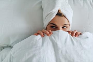 View from above of happy attractive young woman lying down on the bed and awake. Top view of female looking aside covers her face with white blanket in the morning. Copy space for advertising