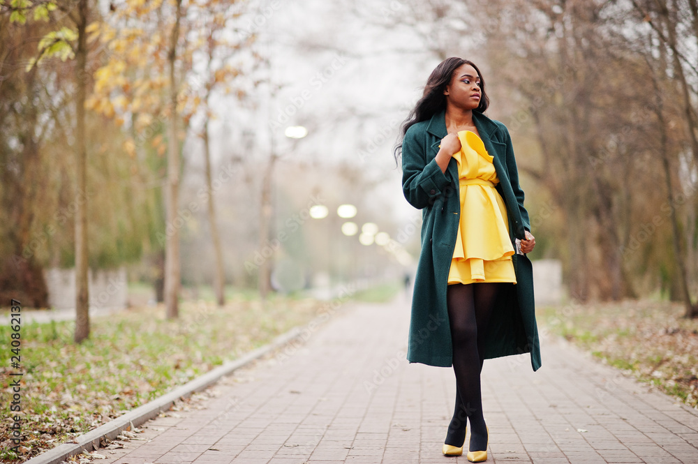 Wall mural stylish african american woman at green coat and yellow dress posed against autumn park.