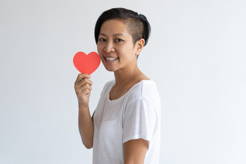 Happy Asian woman holding paper heart. Lady wearing t-shirt and looking at camera. Saint Valentines Day concept. Isolated side view on white background.