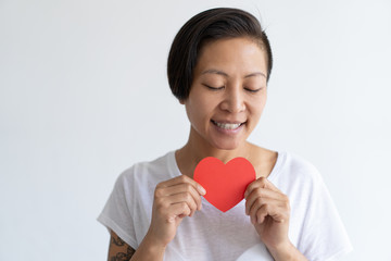 Happy Asian woman posing with paper heart and her eyes closed. Sensitive lady wearing t-shirt. Saint Valentines Day concept. Isolated front view on white background.