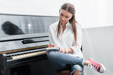 girl with notebook sitting near piano and composing music at home