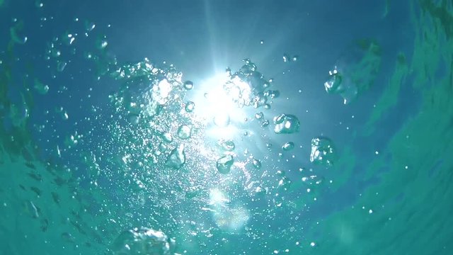 BOTTOM UP, UNDERWATER: Air bubbles floating towards the surface of the tranquil ocean on a sunny day in the Pacific. Blinding summer sunlight shining on the calm turquoise colored sea and big bubbles.