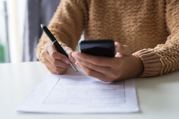 Close-up of lady in sweater checking message on smartphone. Unrecognizable woman filling tax form. Paperwork concept