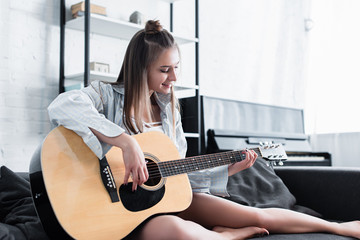 beautiful musician sitting on sofa, smiling and playing acoustic guitar at home