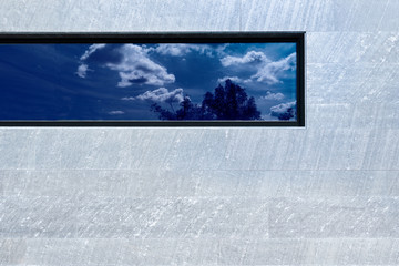 Moderne Fassade aus grauen Steinplatten mit einem länglichen Fenster, in dem sich der Himmel...