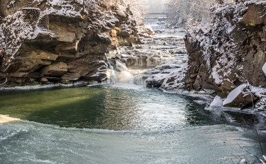 Naklejka premium icy winter waterfall steaming between stones, orange sunlight, frozen pond