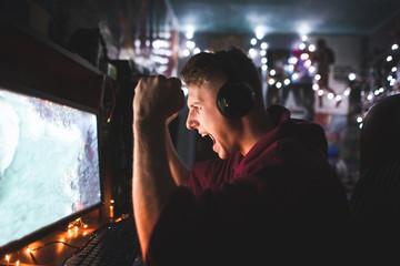 Gamer in his headphones emotionally rejoices in victory,looks at the computer screen. Portrait of an emotional young man playing video games at night on a computer in a cozy room at home and rejoicing
