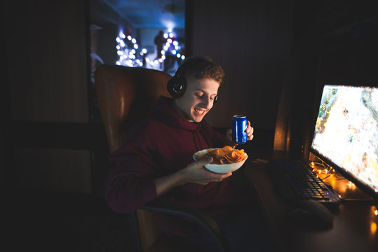 Happy Young Man Sitting At The Work Desk At Home, Looking At The Screen, Smiling And Holding A Can Of Cola. Portrait Of A Positive Gamer Using A Computer, Drinking Cola And Eating Chips At Night.