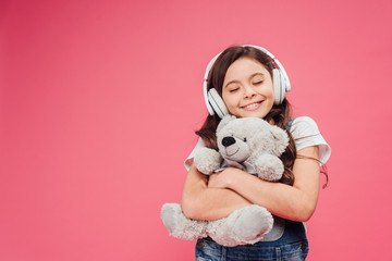 happy kid in headphones hugging teddy bear isolated on pink