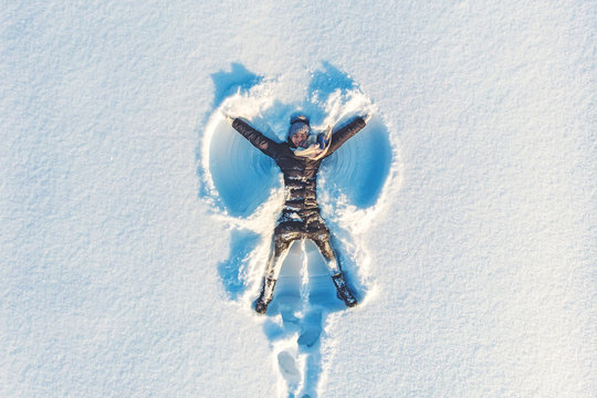 Top aerial view of young happy smiling girl making by arms snow angel figure and lying in snow, winter outdoor activity