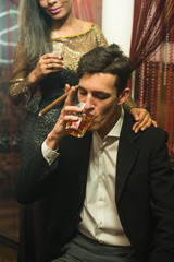 YOung stylish caucasian Man in black suit smoking cigar and drinking whiskey at night in a company with pretty african girl