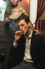 YOung stylish caucasian Man in black suit smoking cigar and drinking whiskey at night in a company with pretty african girl