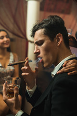Young stylish caucasian Man in black suit smoking cigar and drinking whiskey at night in a company with pretty middle eastern girl