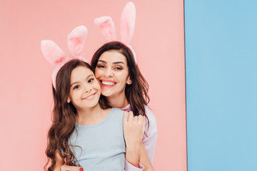 beautiful woman in easter bunny ears hugging child and looking at camera on blue and pink background