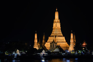 ornate temple, illuminated with warm lights, standing on the edge of a body of water. at Wat Arun,...