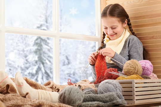 Child girl is sitting on a window sill with wool yarns and knitting. Beautiful view outside the window - sunny day in winter and snow.