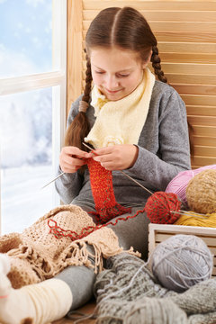 Child girl is sitting on a window sill with wool yarns and knitting. Beautiful view outside the window - sunny day in winter and snow.