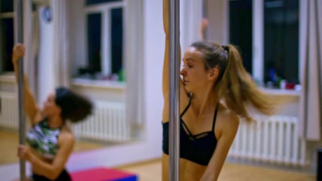 nice slim blond girl with long pony tail in black top practices on pylon at strip pole dance lesson in gym
