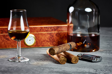 Three Cuban cigars on a stone table with a lighter and a wooden humidor with a glass and a bottle...