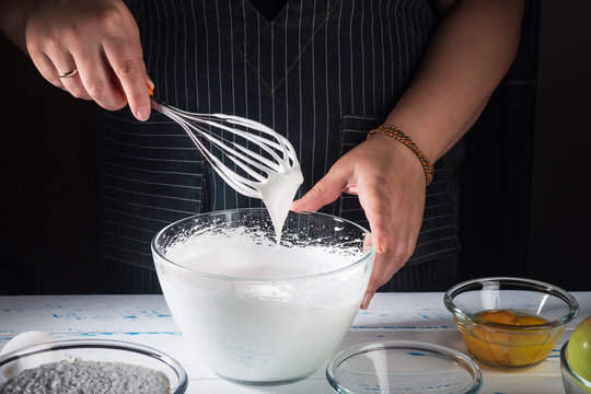 Female Chef Whipping Egg Whites  To Soft Peaks In Glass Bowl With Whisk And Hand On White Wooden Table.  Making Apple Pie - Charlotte.