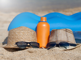 Pair of sunglasses, sunhats and sunscreen protection cream at the sand near the blue air mattress...