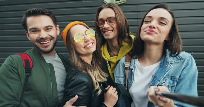 Portrait of the young cheerful male and female friends hipsters laughing and having fun while taking selphie photos with selfie stick. Close up. POV. Outdoor.