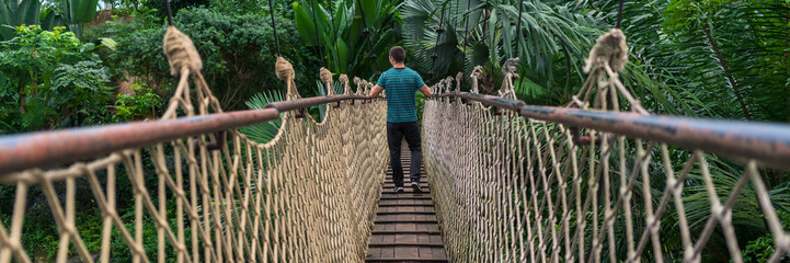 Obraz premium Młody człowiek przechodzi przez wiszący most w panoramie dżungli