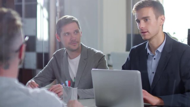 Medium shot of three young businessmen discussing project together during meeting in modern office