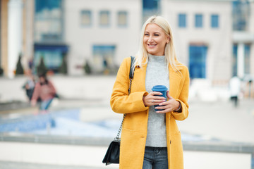 Coffee to go. Beautiful young blond woman in bright yellow coat holding coffee cup and smiling while walking along the street 