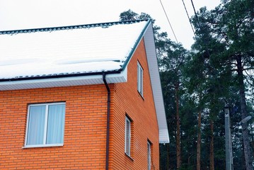 part of a brick red house with windows with white snow on the roof against the background of green pine and sky