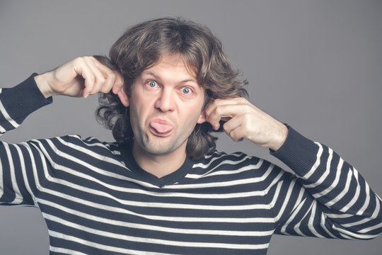 Close up of man making silly monkey face isolated on grey background. Guy grimaces monkey. Funny male holding his ears and shows tongue looks like monkey, pretending chimpanzee, entertaining children