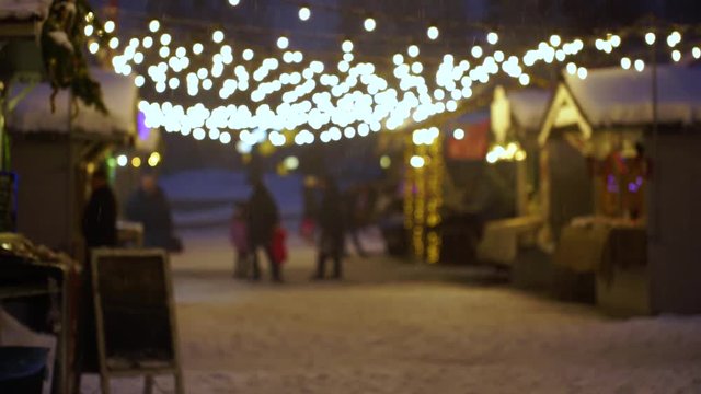 Light holiday blurry bulbs, falling fresh snow and bokeh background of night Christmas time festival city life. People walking. Real time 4k video footage.