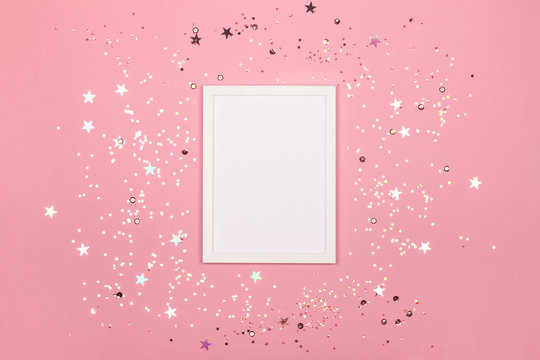 Festive background with blank white photo frame on pink pastel with scattered confetti. Mock up for photo or text. Top view. Flat lay style.