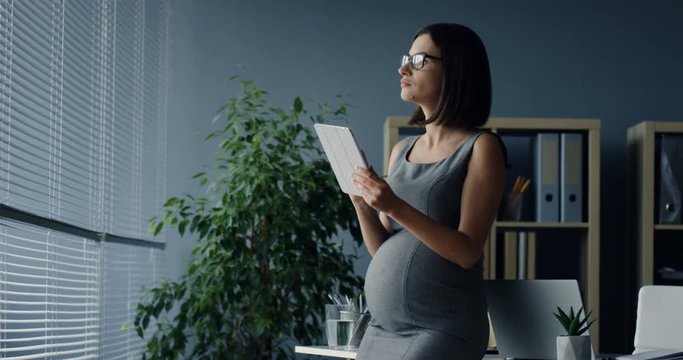 Beautiful Caucasian pregnant woman working while standing in the office, using taplet computer and drinking water.