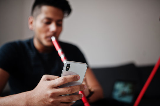 Handsome and fashionable indian man in black sitting at room, smoking hookah and looking on mobile phone.