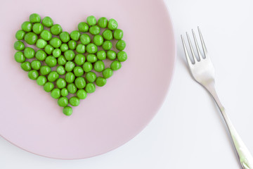Green peas heart shaped, pink plate and a fork on white table. St. Valentine's day concept