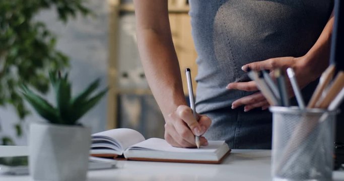 Close up of the hand of the female pregnant woman writing something in the notebook with a pen, making notes. At office.