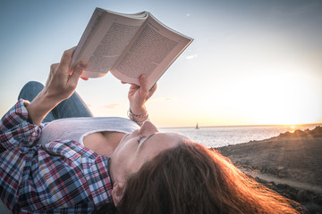 Young redhead woman with plaid shirt and jeans reads a book lying near the ocean Sailing boat on...