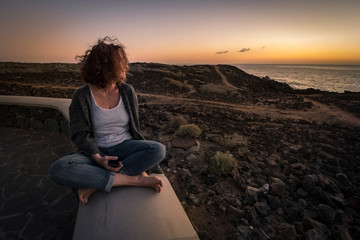 Young woman sitting leg crossed with sunset on the ocean in background. Girl enjoying panorama near the sea with a smartphone in hand. Happy caucasian trendy female with curly hair relaxing outdoor 