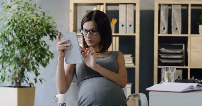Pregnant attractive woman office worker scrolling and tapping on the tablet computer with a smile at her workplace.
