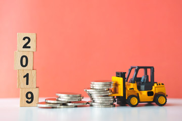Miniature forklift vehicle working on stack coins on wooden block year 2019 background using as business and industry concept