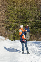 Mother with son in arms and backpack stands against the background of coniferous forest and snow-covered road. Winter day.