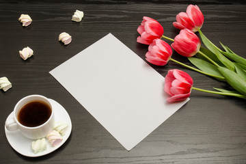 Fototapeta na wymiar Holiday concept. Bouquet of pink tulips, a cup of coffee, marshmallow and sheet of paper on a black wooden background