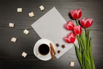Obraz na płótnie Canvas Holiday concept. Bouquet of pink tulips, a cup of coffee, marshmallow and sheet of paper on a black wooden background. Directly above.