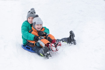 Fototapeta na wymiar Two joyful boys slides down the hill on snow saucer. Brotherly friendship. Winter day