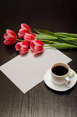 Holiday concept. Bouquet of pink tulips, a cup of coffee and sheet of paper on a black wooden background. Vertical frame