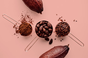 Rose gold measuring cups of cocoa beans, cacao nips, cocoa powder and cocoa pods on a pink...